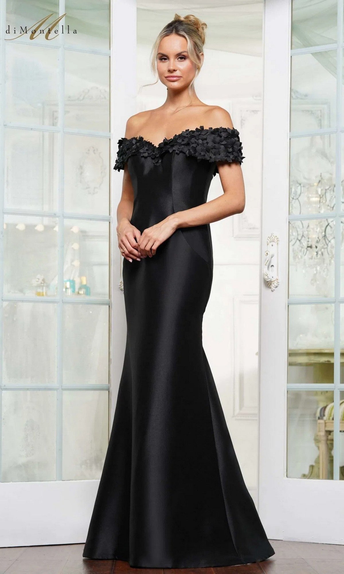 A woman stands indoors wearing the Rina Di Montella RD3254, a black, floor-length, off-the-shoulder formal gown with floral detailing on the neckline and styled in an elegant mermaid silhouette.